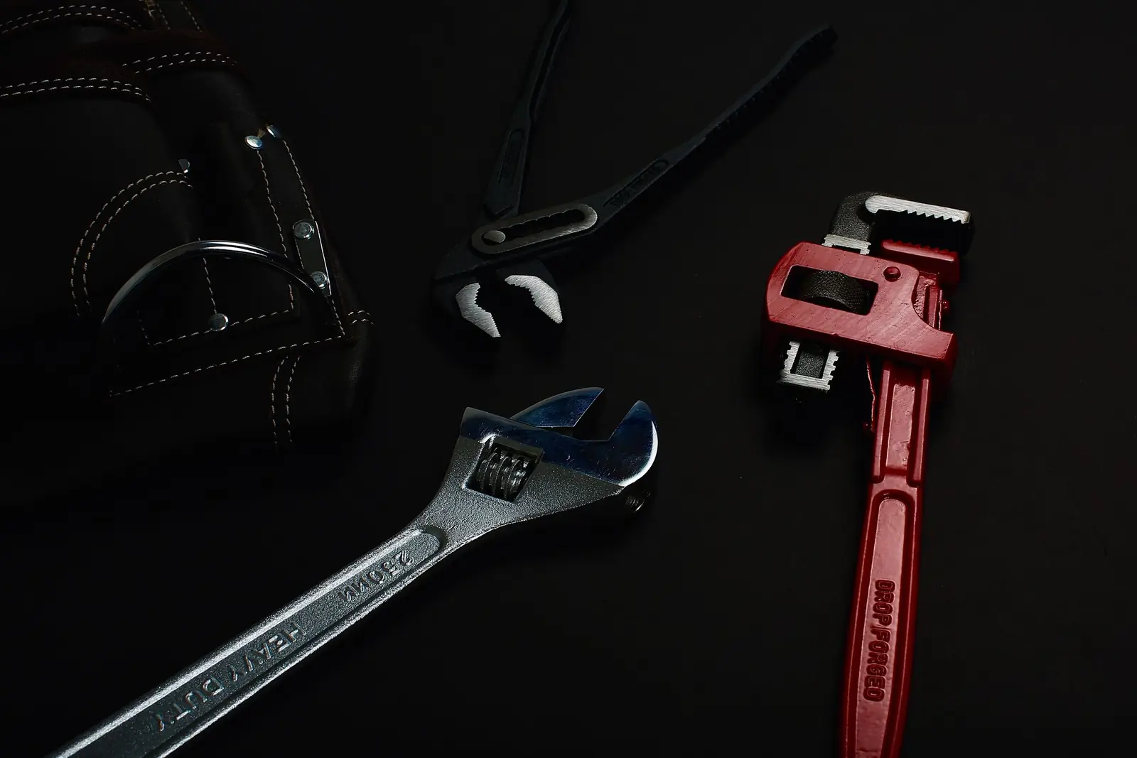 an adjustable wrench, a red pipe wrench, water pump pliers and a leather tool pouch laid out on a dark surface