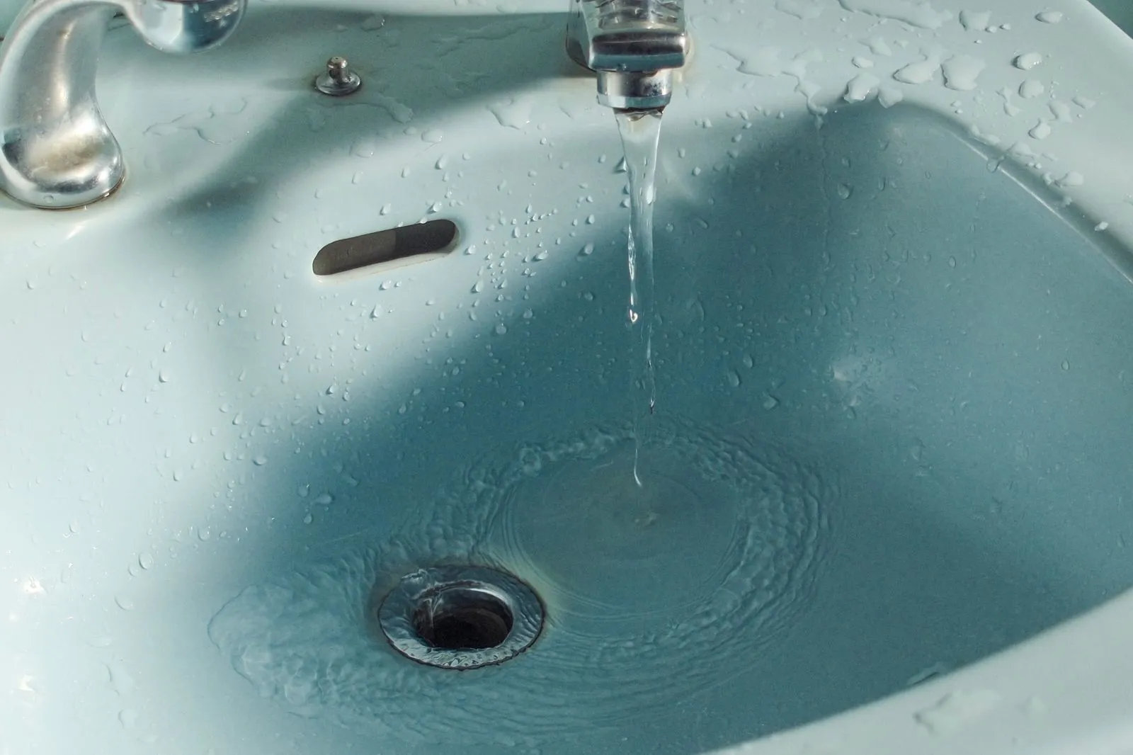 water pooling around the waste of a pale blue bathroom sink with the tap running and the overflow slot above it